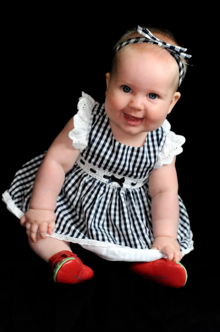 A smiling baby girl in a black-and-white gingham dress and red shoes against a black backdrop.