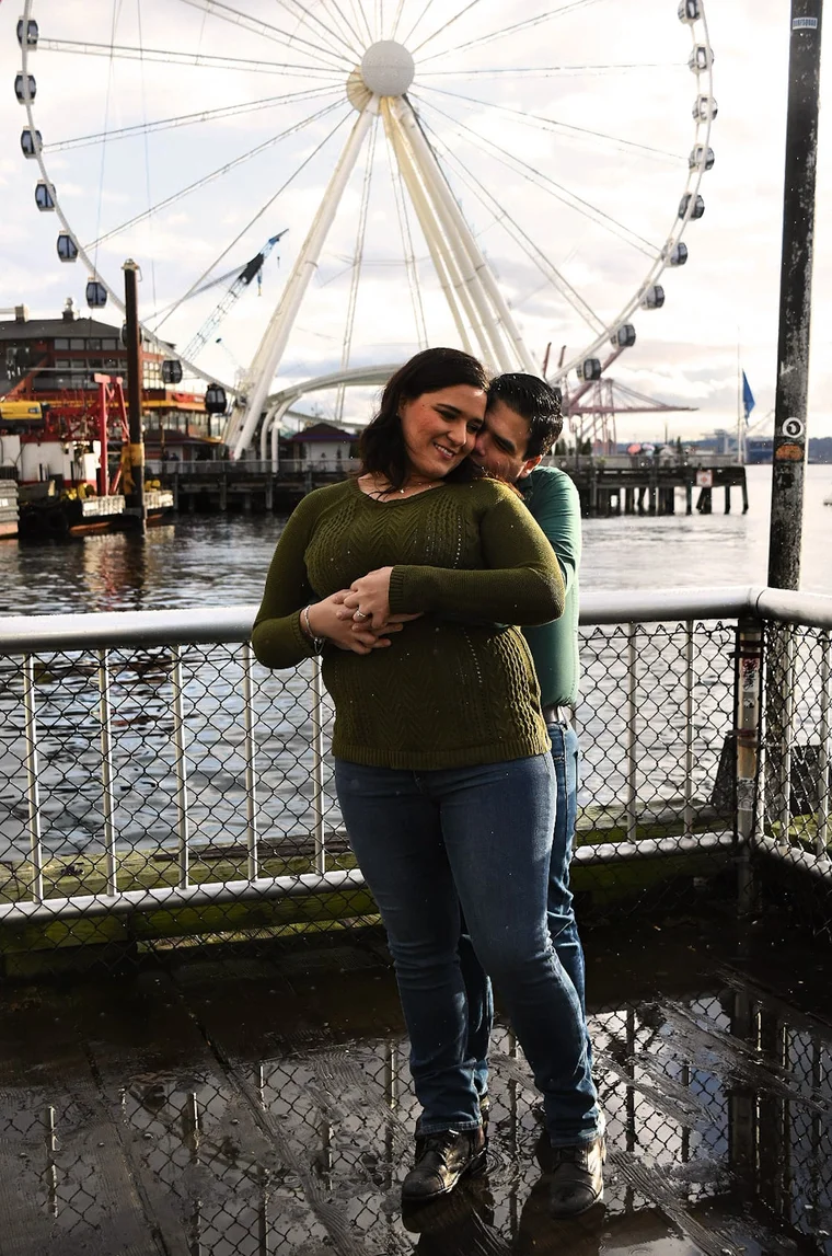 A couple embracing on a waterfront pier with a Ferris wheel rising behind them.