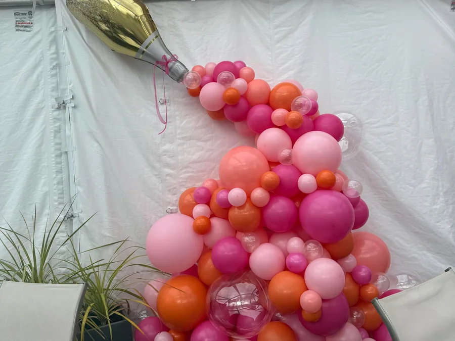 Pink and orange balloon arrangement on a white backdrop with a champagne bottle on top