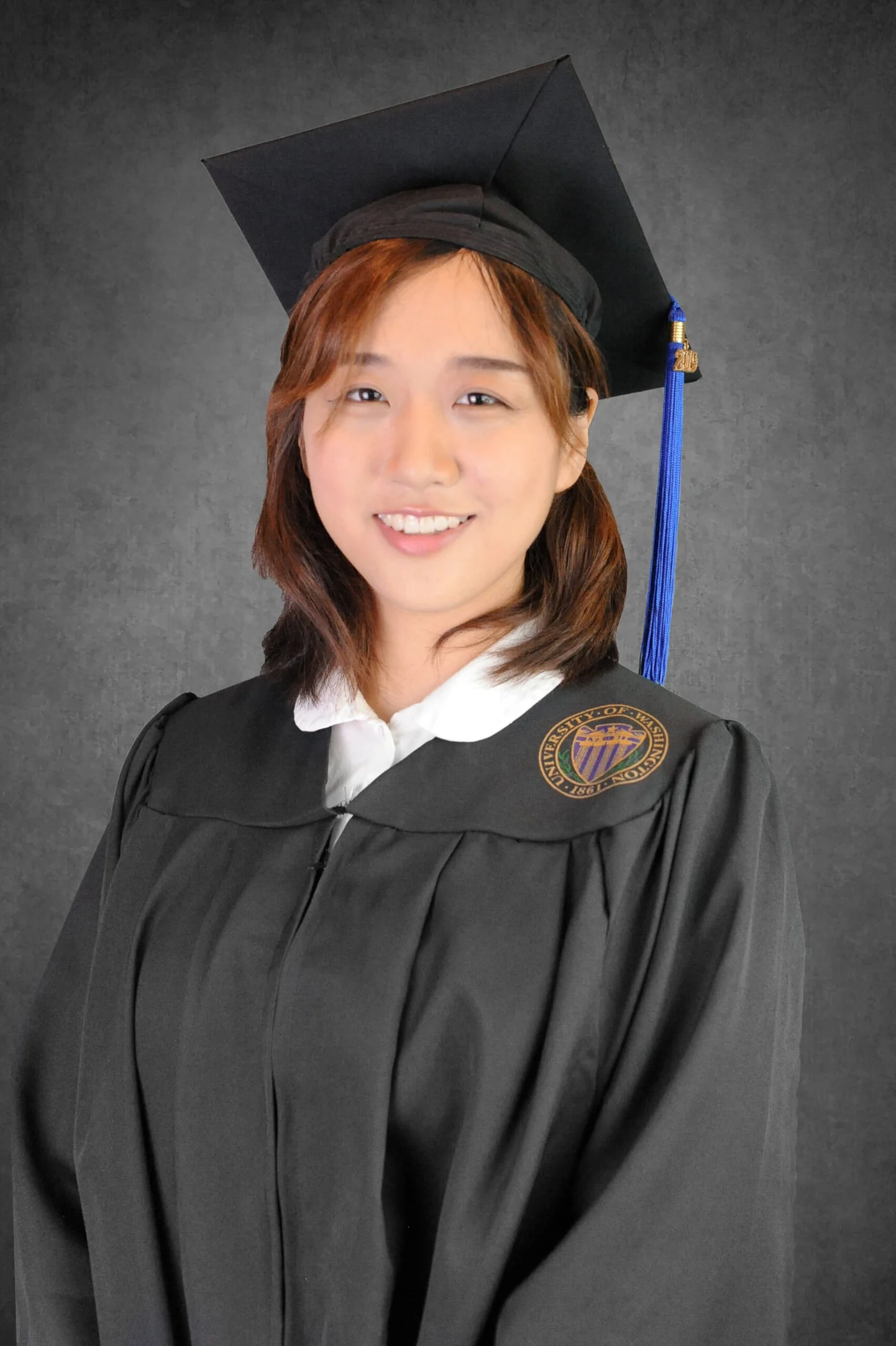A smiling graduate in a cap and black gown, photographed against a grey studio backdrop.