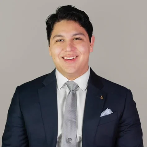Portrait of Aaron Peña-Diamond, smiling in a navy suit with a grey tie and pocket square.