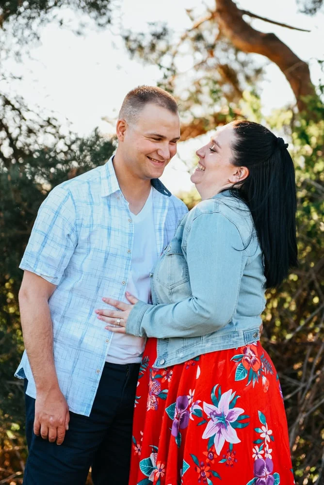 Smiling couple in denim and floral red skirt embracing among sunlit trees