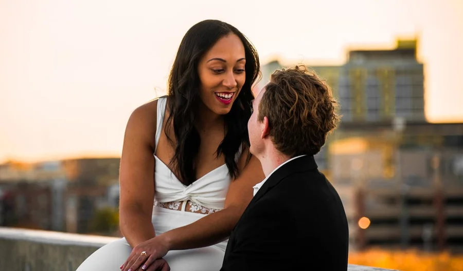 Smiling engaged couple sitting close on a railing with a sunset skyline behind