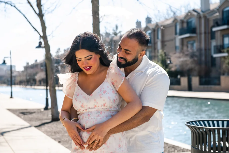 Expecting couple in white embracing a baby bump on a waterfront path