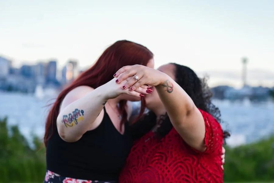 Two women embracing with tattooed arms in front of a city skyline across the water