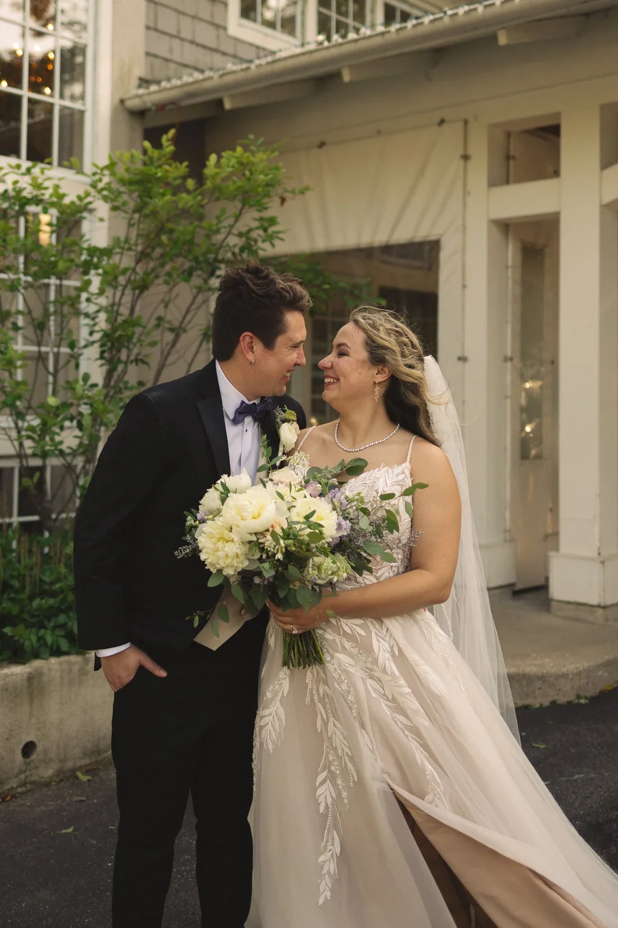 Bride and groom smiling together holding a white bouquet outside a lakeside clubhouse