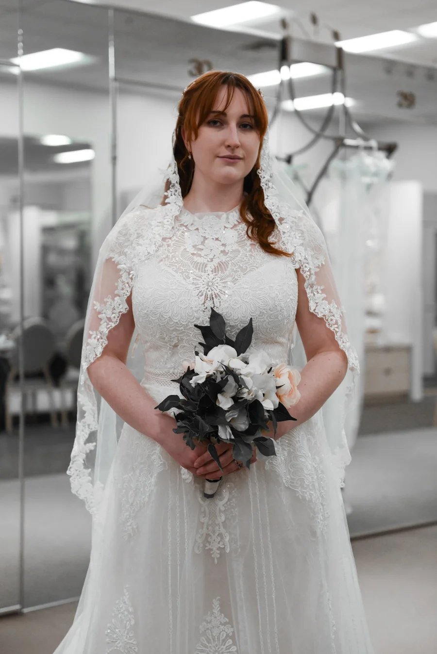 Bride in a lace gown and veil holding a dark bouquet in a bridal shop