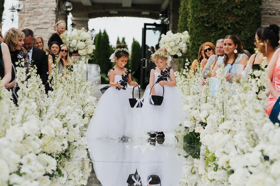 Two flower girls in white dresses walking down an aisle bordered by white blooms