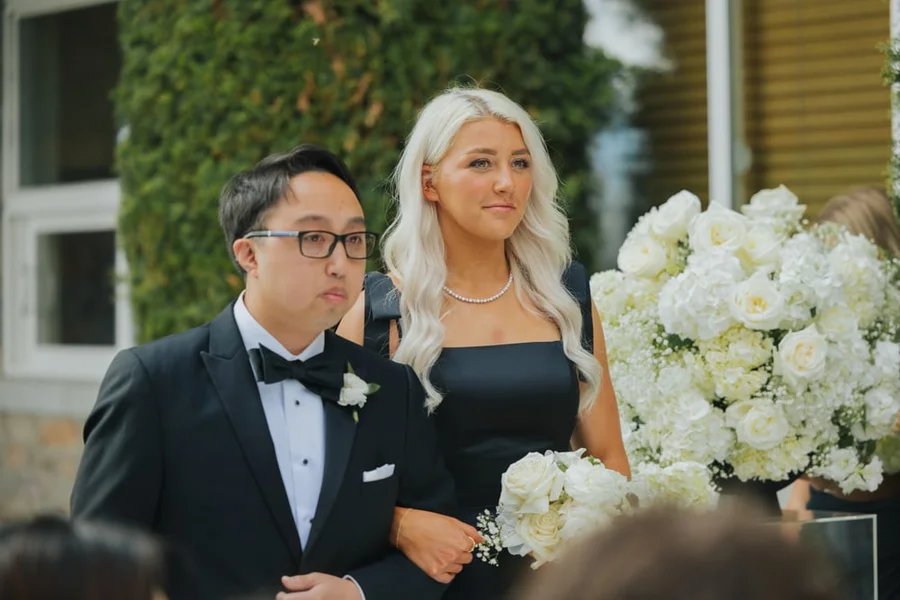 Groom in a tuxedo with a bow tie and bride standing among white flowers