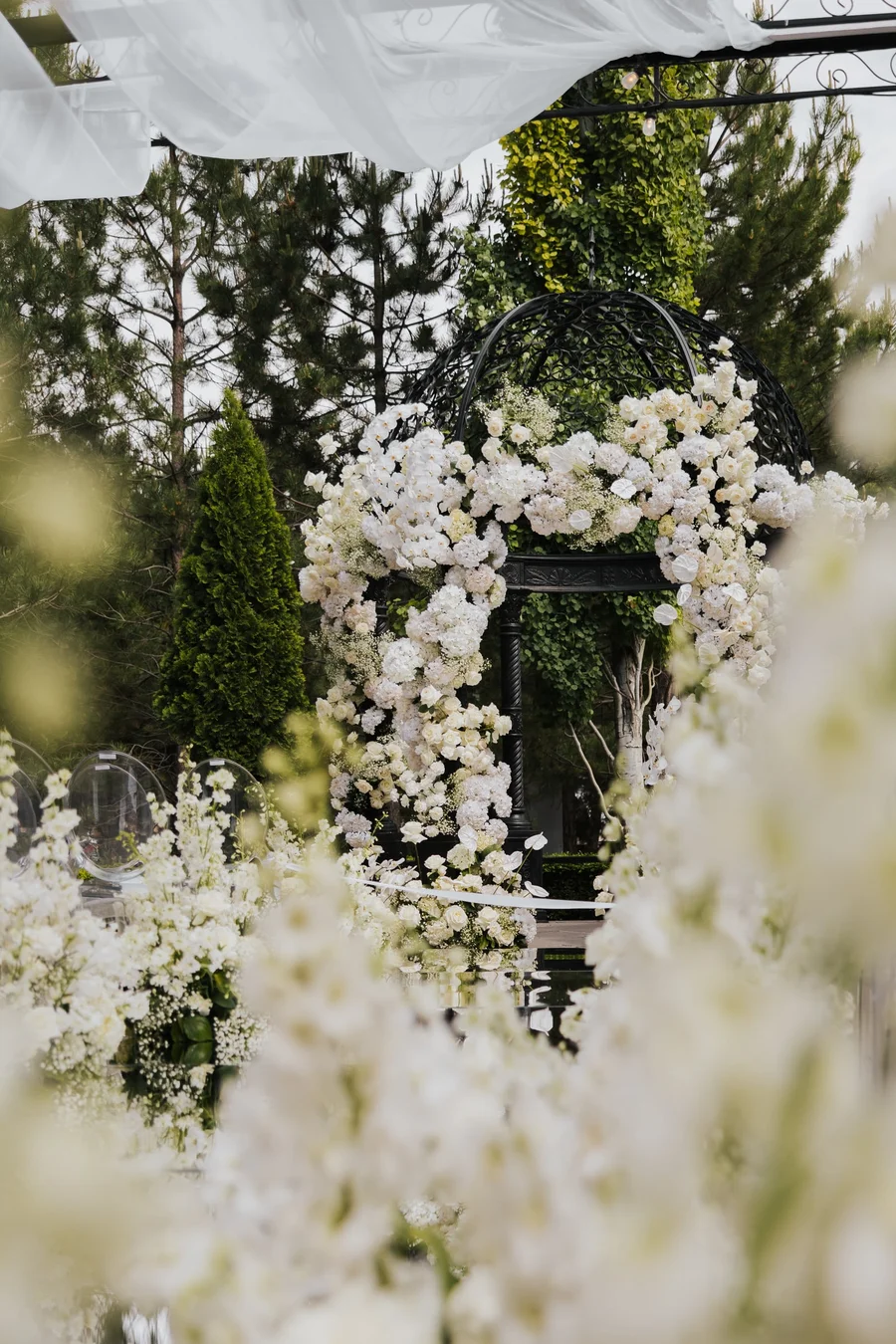 Tall white floral arch framed by trees at an outdoor ceremony
