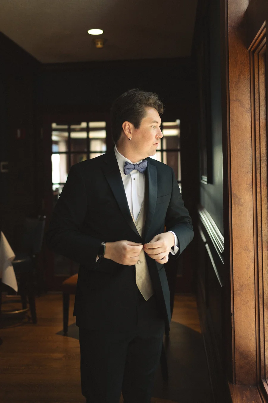 Groom in a dark suit and bow tie adjusting his jacket by a window