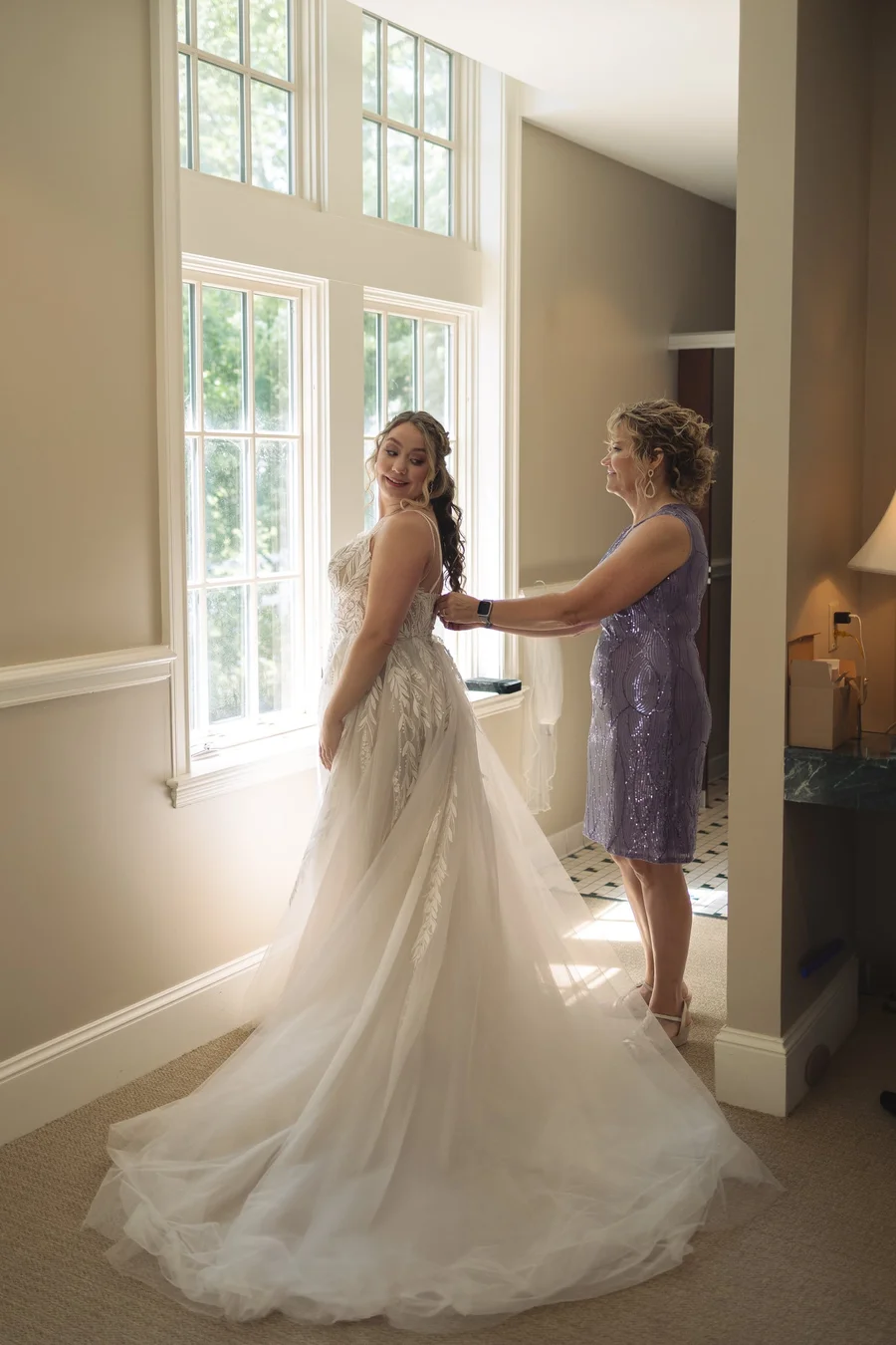 Bride in a long gown with veil being assisted by a woman in lavender