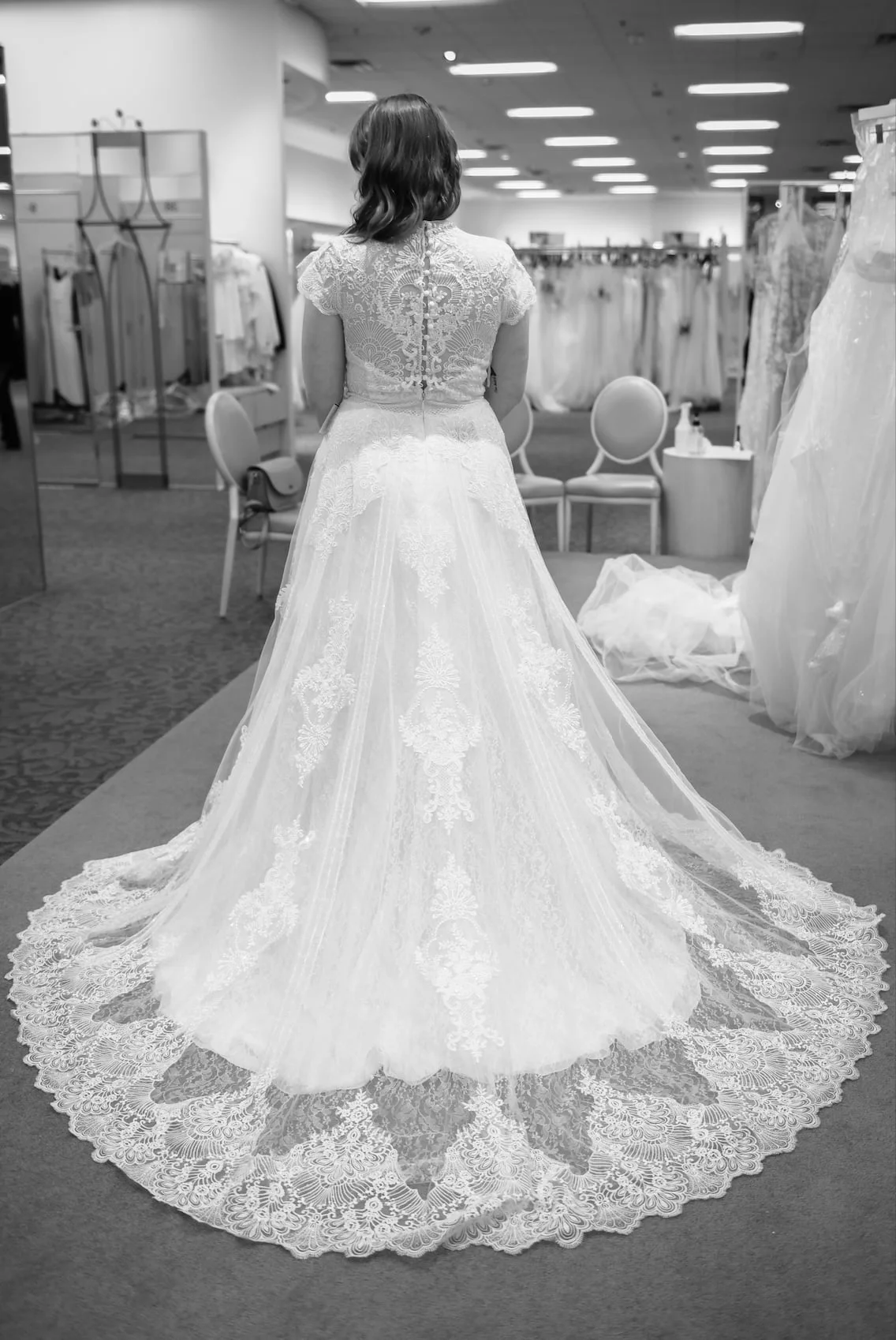 A bride seen from behind in a lace gown with a long train, standing in a bridal salon.