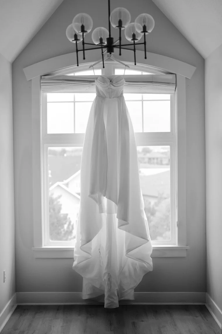A wedding gown hanging in a tall arched window beneath a black chandelier, in black and white.
