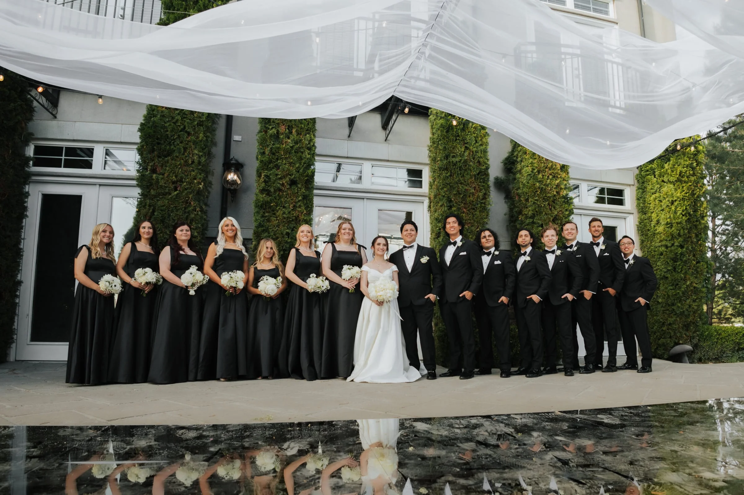 A wedding party in black formalwear lined up on the terrace of an estate, with a sheer white canopy overhead.
